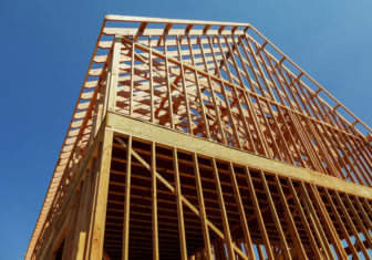 A single family home under construction. The house has been framed and covered in plywood. Stacks of board timber in front and stack of 2x4 boards on the top.